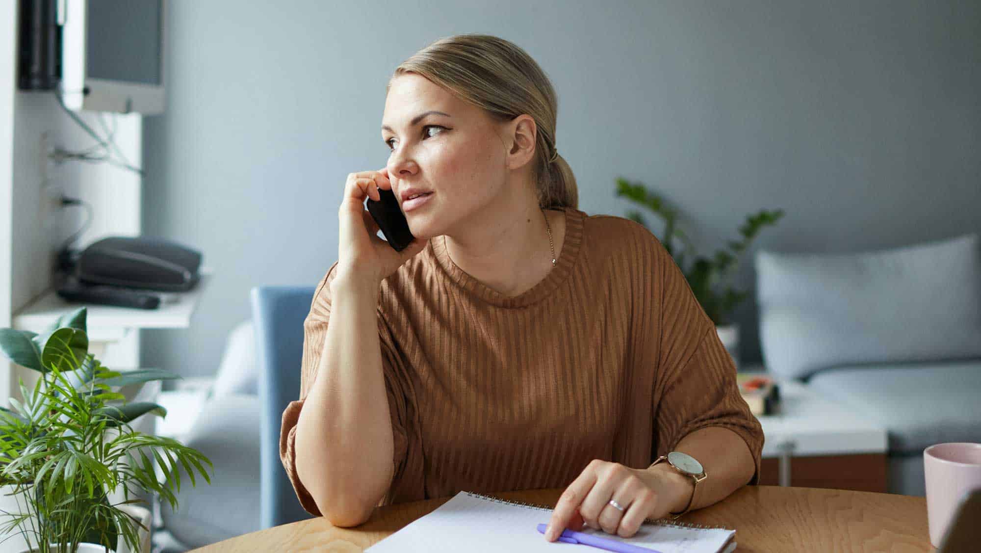 A woman on the phone at her kitchen table, making notes